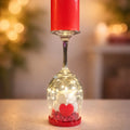 Decorative wine glass with a red candle, small figurine, and heart-shaped object on a blurred background.